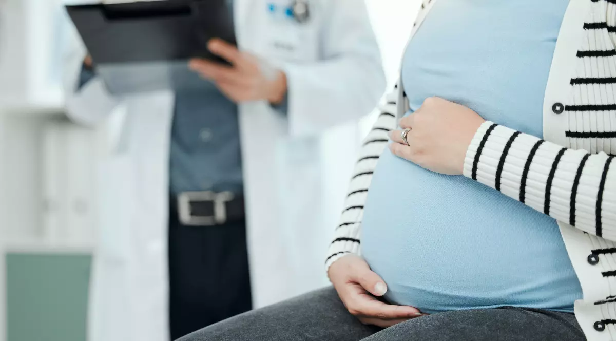 O femeie însărcinată își ține mâna pe burtă, în timp ce un medic se află în fundal, analizând documente. Imaginea sugerează un control medical și o situație care necesită atenție și îngrijire de specialitate.