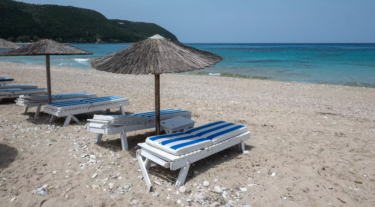 O plajă liniștită, cu șezlonguri albe și umbrele din stuf, se întinde de-a lungul unei ape turcoaz limpezi. În fundal, dealurile verzi completează peisajul mediteranean relaxant.