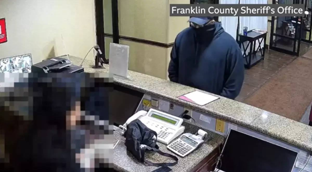 Un bărbat îmbrăcat în negru, mascat, cu șapcă în cap, care stă în recepția unui hotel și cere bani unei funcționare