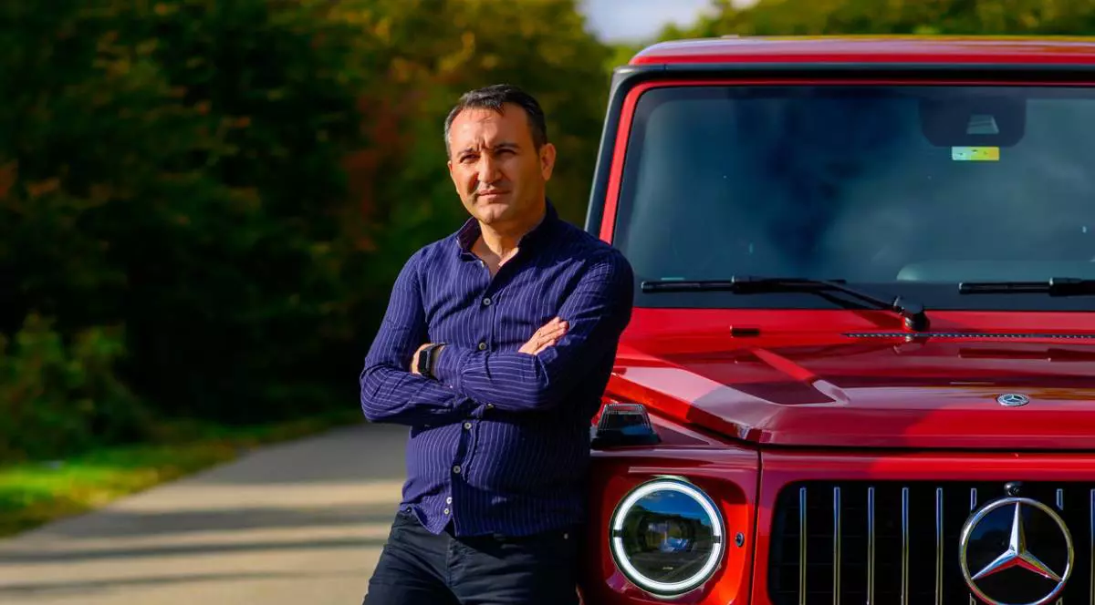 Un barbat in camasa bleumarin si blugi inchisi la culoare, Ibrahim Günbeyi, sprijinit de o masina rosie de teren in mijlocul unui drum mărginit de padure