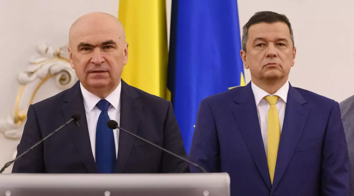 Ilie Bolojan, în stânga, îmbrăcat cu un sacou negru, cămașă albă și cravată albastră, vorbind la un pupitru cu un microfon în față. În dreapta, Sorin Grindeanu, îmbrăcat cu un sacou bleumarin, cămașă albă și cravată galbenă.