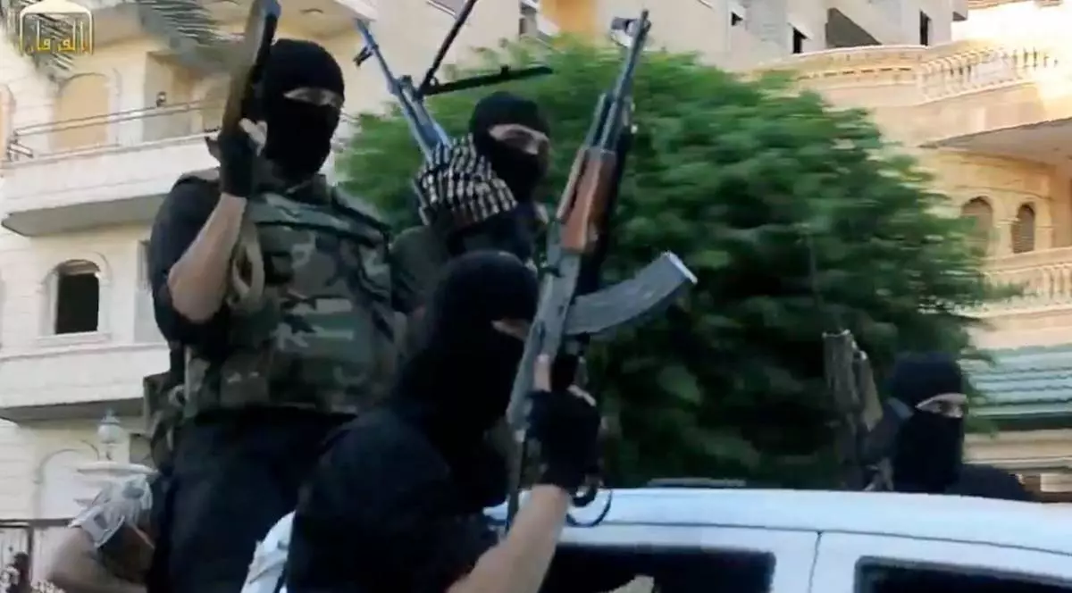 Mai mulți bărbați mascați, înarmați, stau pe și lângă o mașină albă, pe o stradă cu clădiri în fundal.