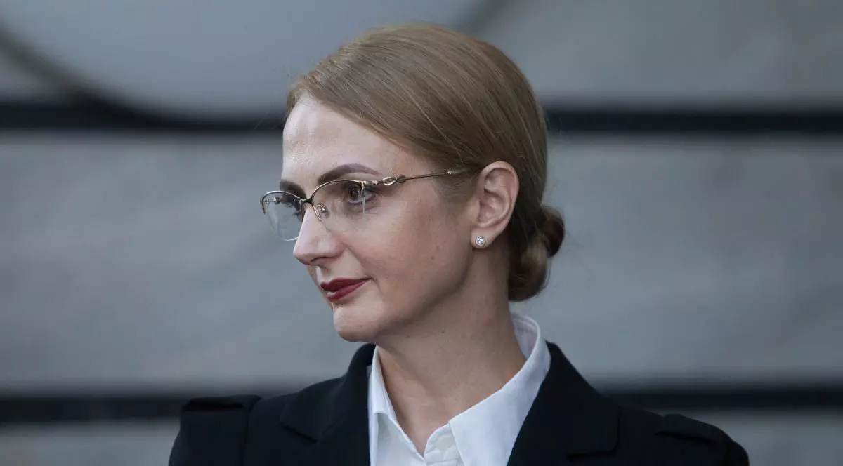 Lia Savonea, cu ochelari de vedere, îmbrăcată cu o cămașă albă și sacou negru, iar pe fundal peretele gri al unei clădiri