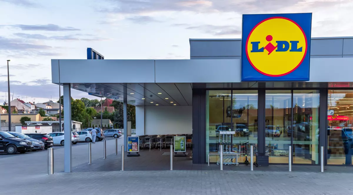 Un magazin Lidl modern este surprins din exterior, cu intrarea largă și logo-ul vizibil deasupra ușilor automate. În fața clădirii se află parcarea și zona pentru cărucioare, sugerând acces facil pentru clienți.