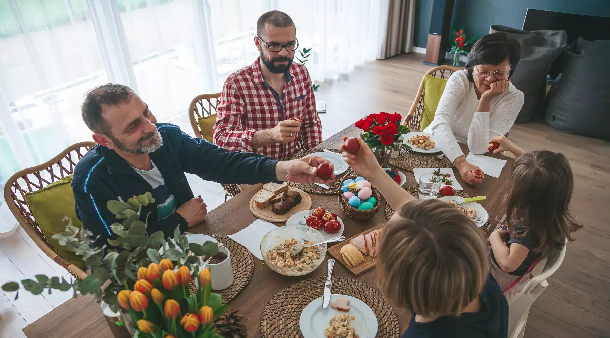 Imaginea surprinde o masă de familie, unde mai multe persoane stau în jurul unei mese, sugerând un moment de reuniune. În fundal se observă un spațiu exterior cu grădini și case, creând o atmosferă de viață cotidiană.