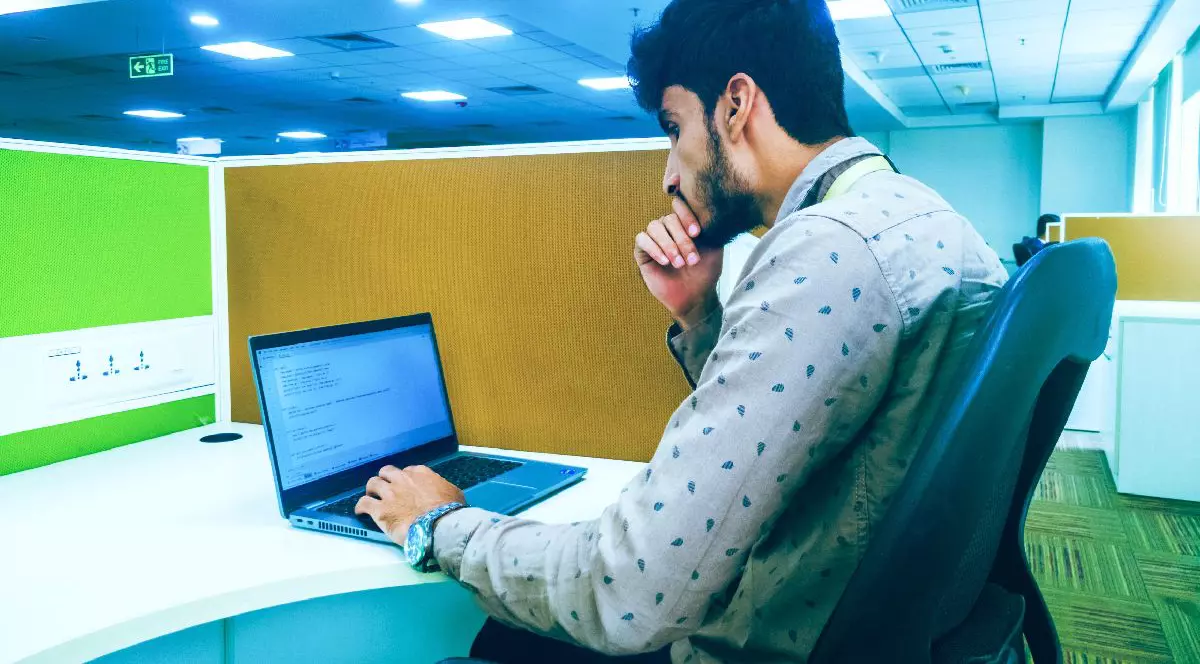 Imaginea surprinde un tânăr concentrat la locul de muncă dintr-un birou, stând la un computer.