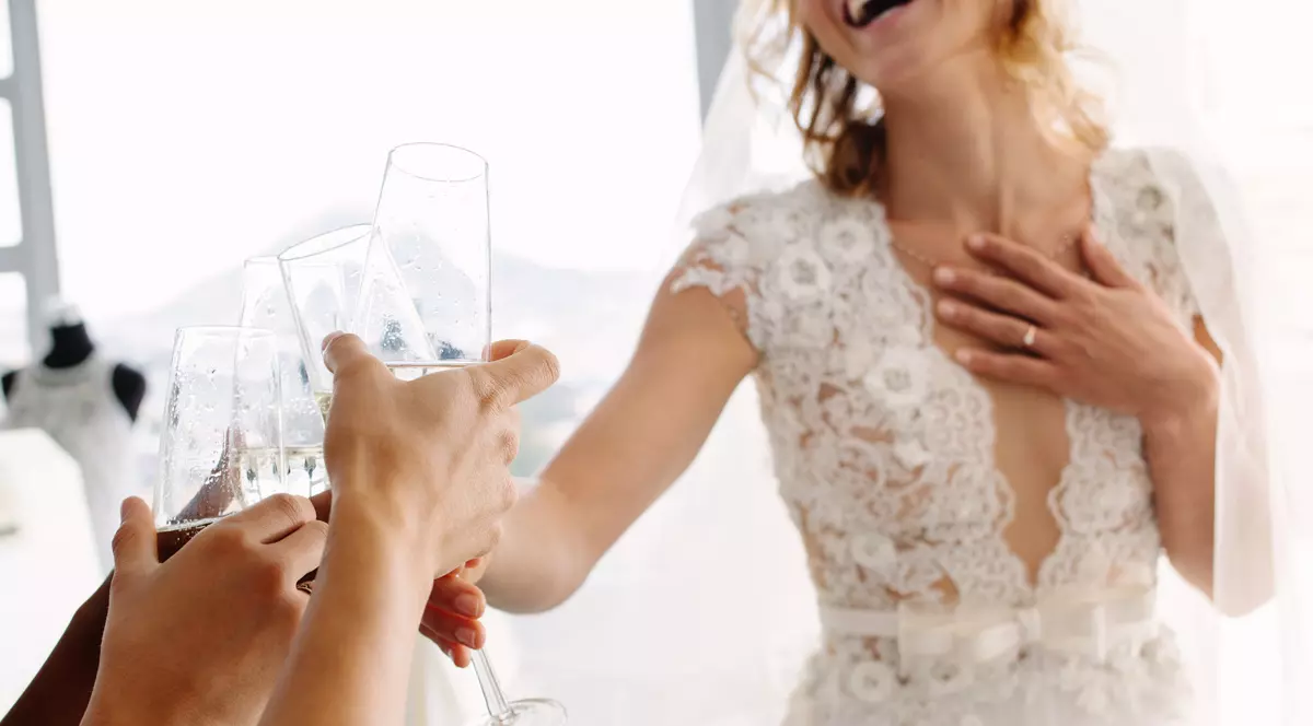 O femeie blondă îmbrăcată în rochie de mireasă ciocnește un pahar cu o altă persoană căreia i se vede doar mâna