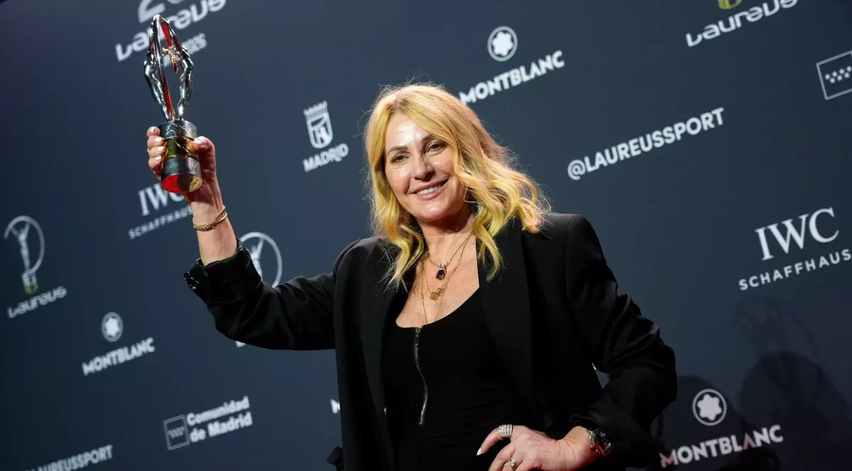 Nadia Comăneci apare zâmbitoare pe covorul roșu, ținând în mână trofeul primit la Gala Laureus, într-o ținută elegantă de culoare neagră. În fundal se observă panoul oficial al evenimentului, cu logo-urile sponsorilor și ale galei.