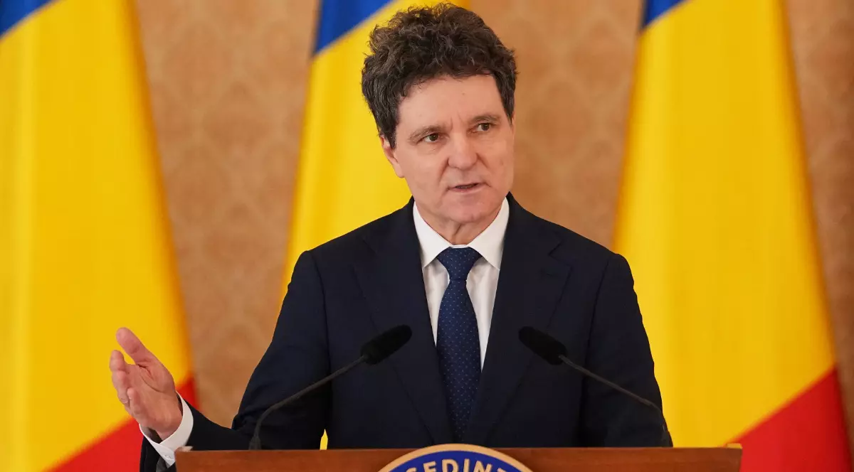 Un bărbat îmbrăcat în costum închis la culoare vorbește la un pupitru oficial, având în față două microfoane. În fundal se văd steagurile României, iar postura lui sugerează că susține un discurs sau o declarație publică.