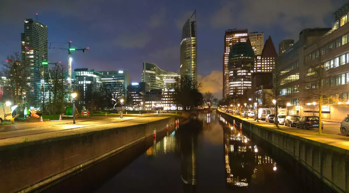 Imaginea surprinde o panoramă nocturnă a orașului Haga, unde clădiri iluminate și zgârie-nori se oglindesc în apele liniștite ale unui canal mărginit de alei pietonale.