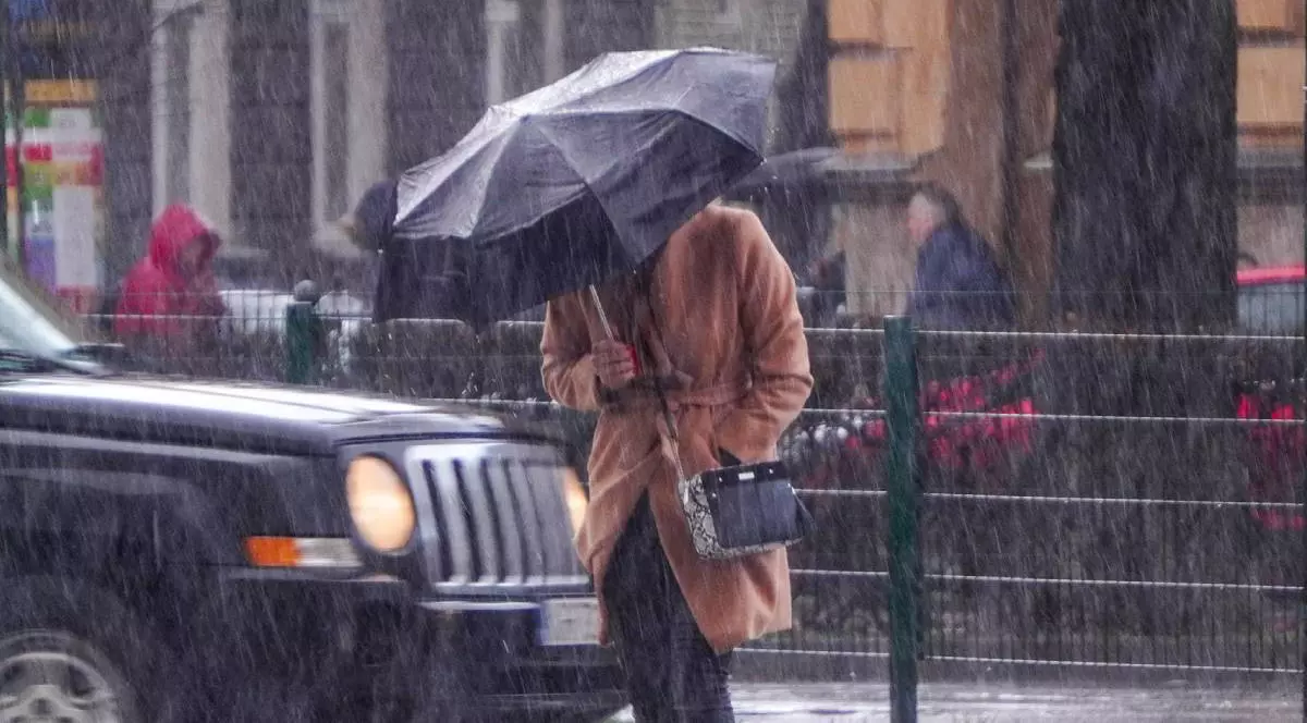 Imaginea surprinde o persoană văzută din fata, cu un palton crem, care se protejează de vant si ploaie sub o umbrelă neagra, într-un cadru urban, se vede o masina neagra cu farurile aprinse, mai multe persoane cu pelerine de ploaie in spate, ploua torential