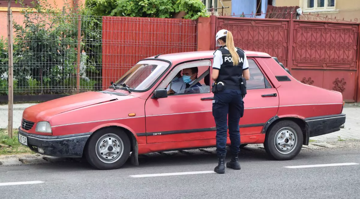 O polițistă îmbrăcată în uniformă controlează actele unui șofer care conduce o Dacia 1300 de culoare roșie