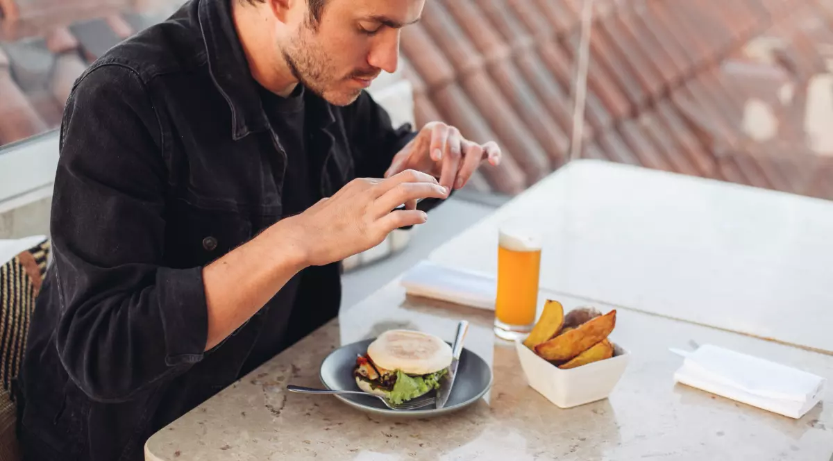 Un bărbat stă la o masă și fotografiază un burger cu telefonul mobil. În fața lui se află și o porție de cartofi și un pahar cu băutură, iar scena sugerează că verifică sau documentează preparatul înainte de a-l consuma.