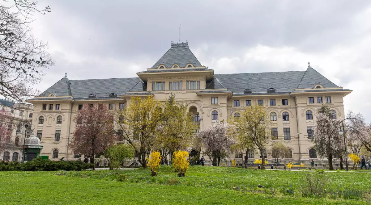 Clădirea Primăriei Municipiului București, istorică, văzută din față, cu o fațadă deschisă la culoare și acoperiș înalt, în stil clasic. În față se află un parc cu iarbă verde, copaci înfloriți și arbuști galbeni. Cerul este acoperit de nori, iar în jurul clădirii se văd câțiva oameni care trec pe trotuar.