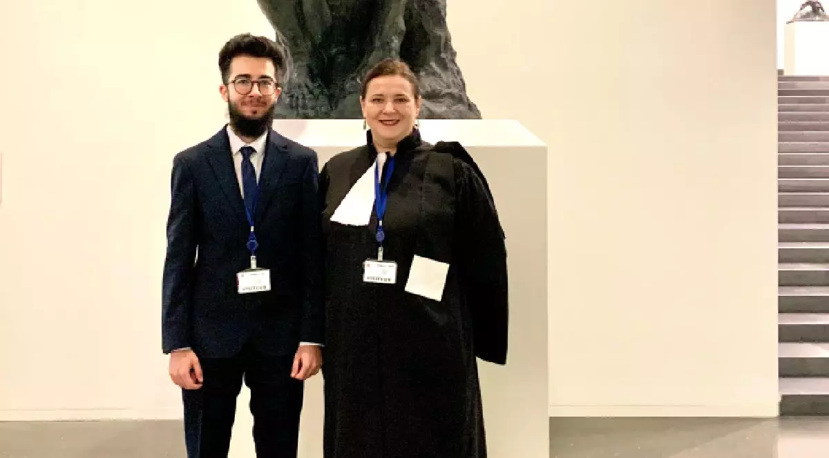 Un tânăr bărbat în costum, cu barbă, alături de o femeie în robă de culoare neagră, de avocat