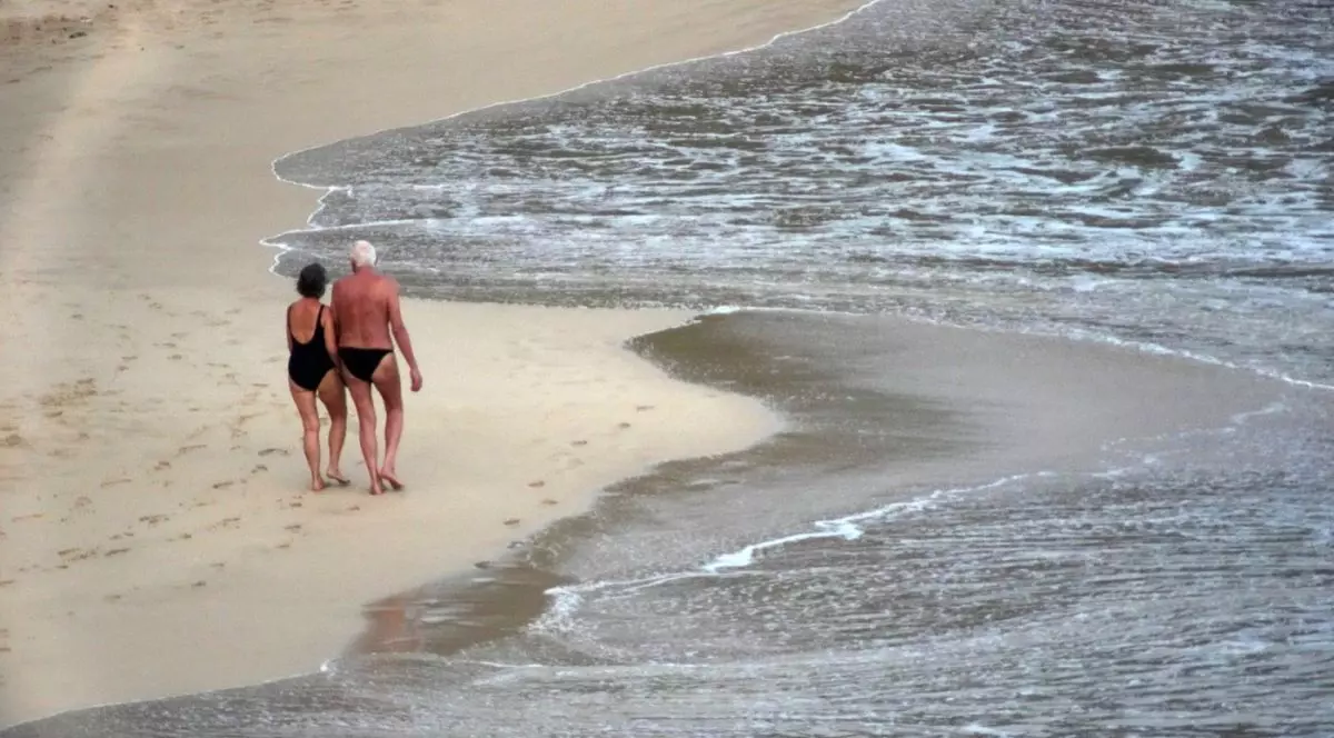 Un cuplu de pensionari în costume de baie se plimbă prin nisipul de la marginea mării, decembrie 2025