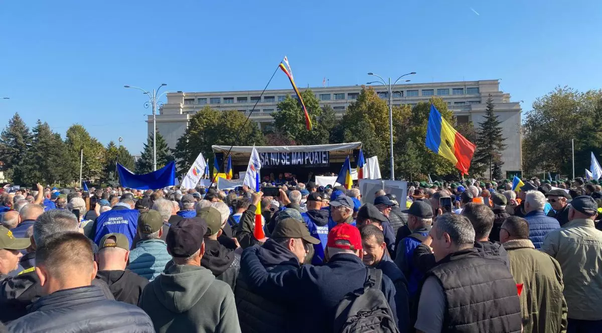 Sute de oameni purtând bannere și steaguri participă la un protest în Piața Victoriei din București, martie 2025