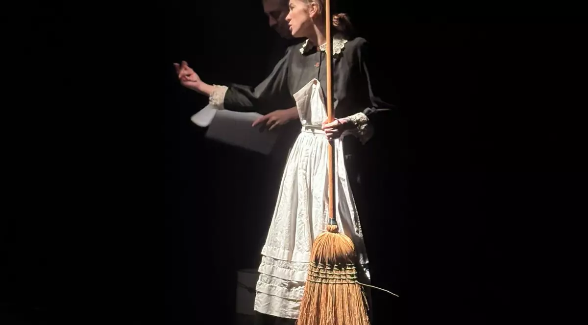Imaginea surprinde o actriță pe scenă, îmbrăcată în costum de cameristă, ținând o mătură și gesticulând expresiv într-o lumină dramatică. Decorul întunecat și jocul de lumini pun accent pe interpretarea sa, sugerând o scenă intensă și teatrală.