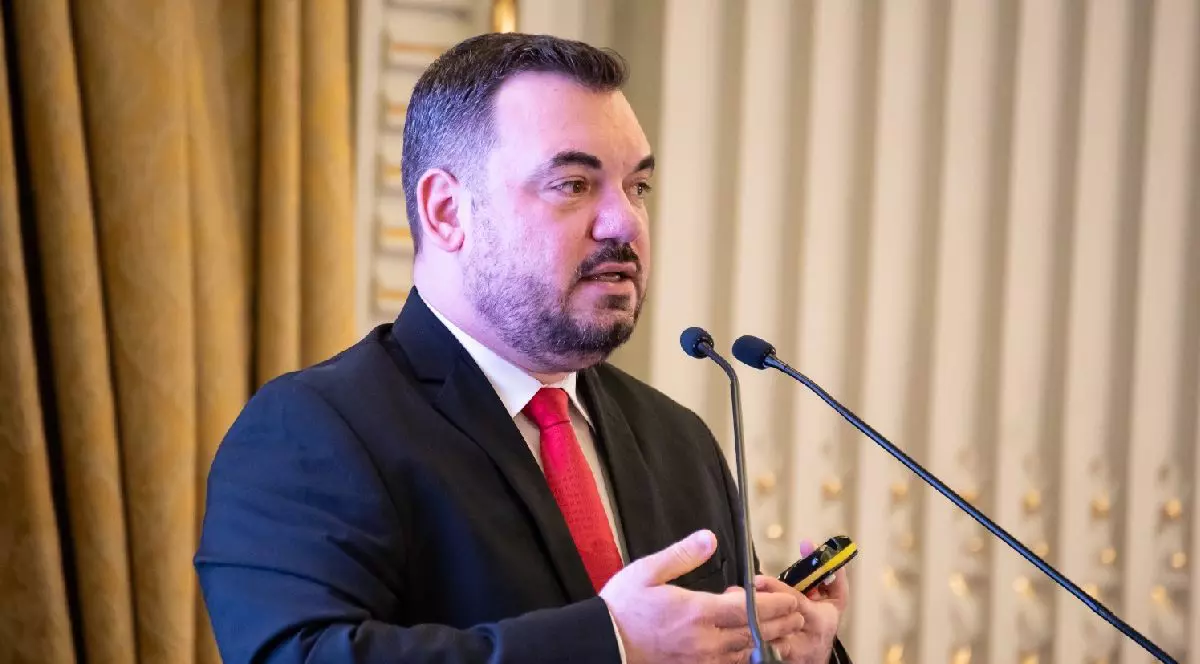 Un barbat in costum cu cravata rosie vorbind la microfon. Razvan Prisada ANMDM despre studii clinice, explica zonele moarte ale cercetarii medicale