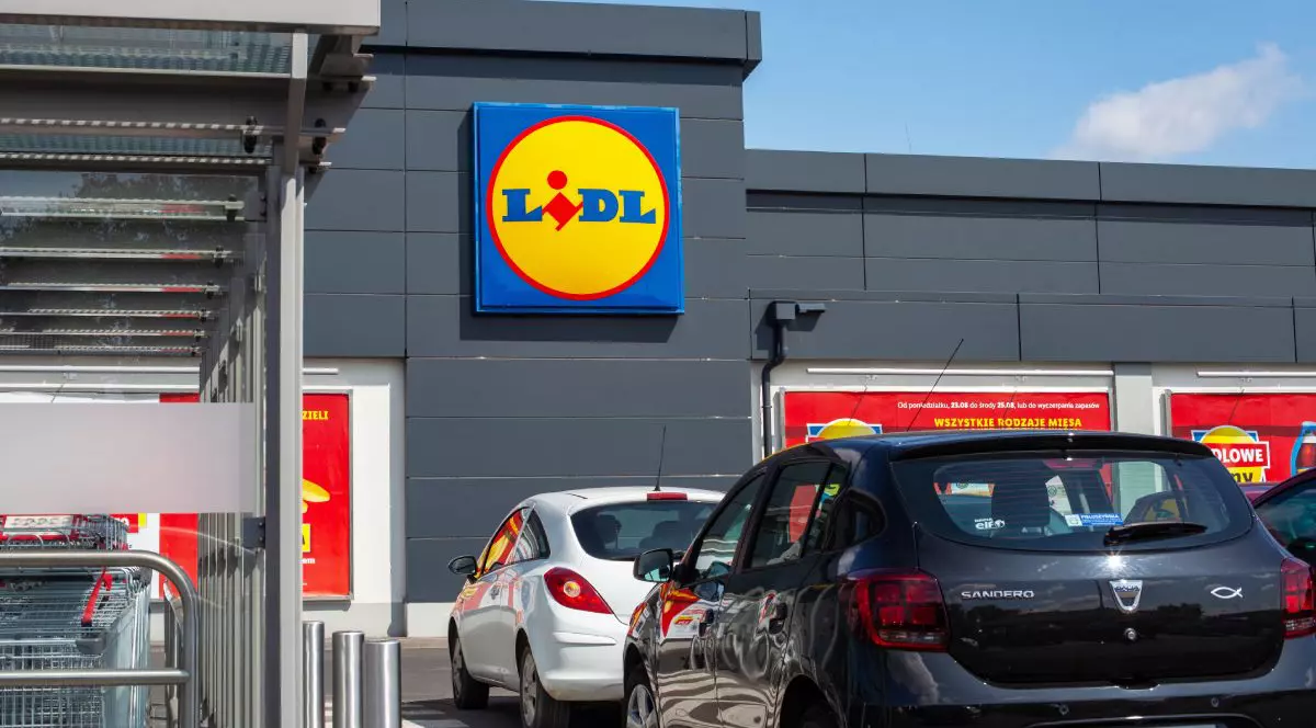 Imaginea prezintă exteriorul modern al unui magazin Lidl, evidențiind logo-ul companiei pe fațada gri a clădirii și câteva autoturisme parcate în față sub un cer senin.