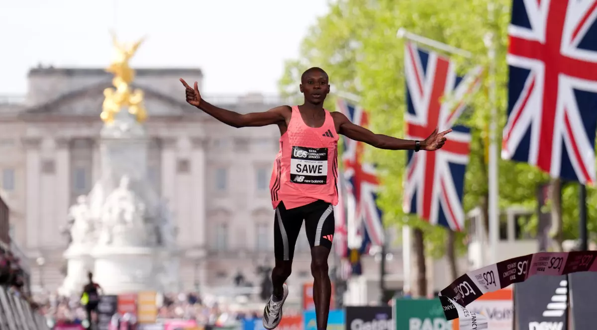 Un alergător kenyan trece linia de sosire cu brațele ridicate, celebrând victoria la maraton. În fundal se văd steaguri britanice și un reper iconic din Londra, sugerând atmosfera unei curse importante.