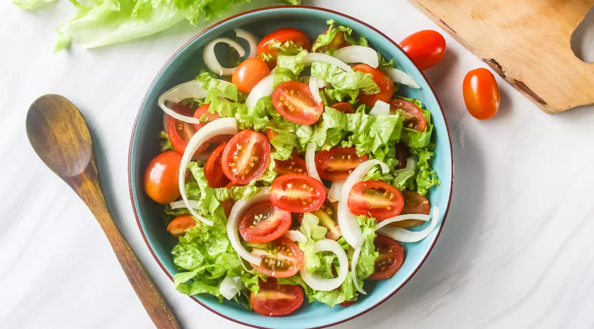 Imaginea prezintă o salată proaspătă, colorată, alcătuită din frunze de salată verde, roșii cherry și felii subțiri de ceapă. Preparatul este așezat într-un bol, lângă ingrediente proaspete și o lingură de lemn, sugerând un stil de viață sănătos.