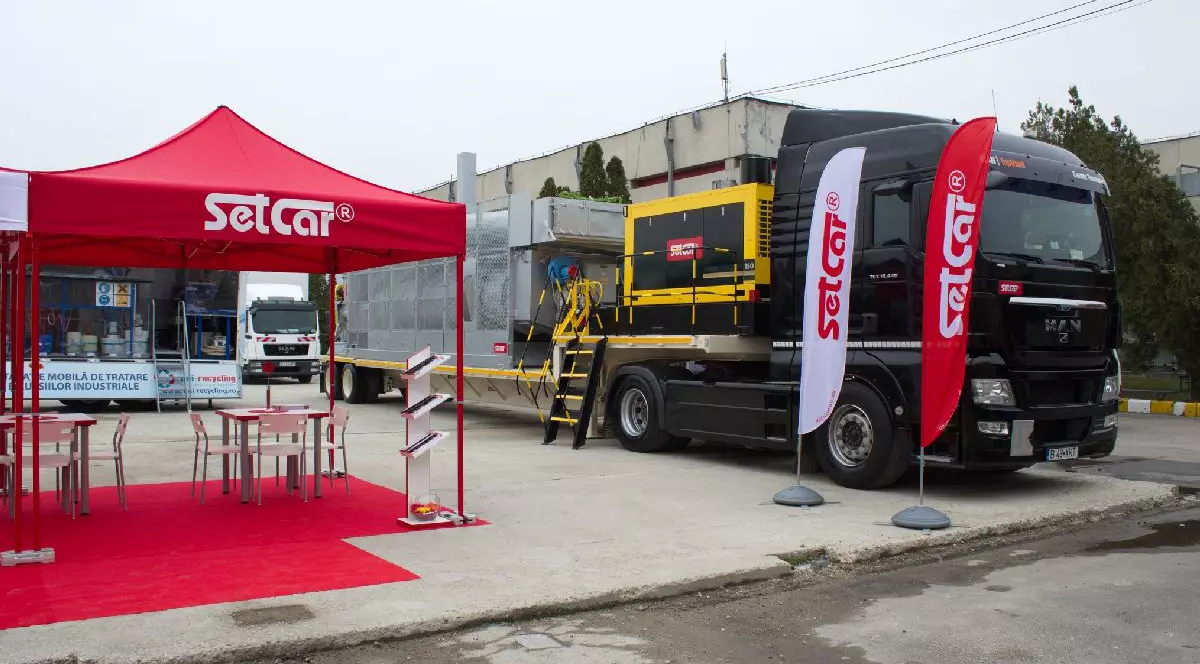 Imaginea prezintă un stand expozițional în aer liber al companiei SetCar, incluzând un pavilion roșu cu mese și scaune lângă un camion de mari dimensiuni.