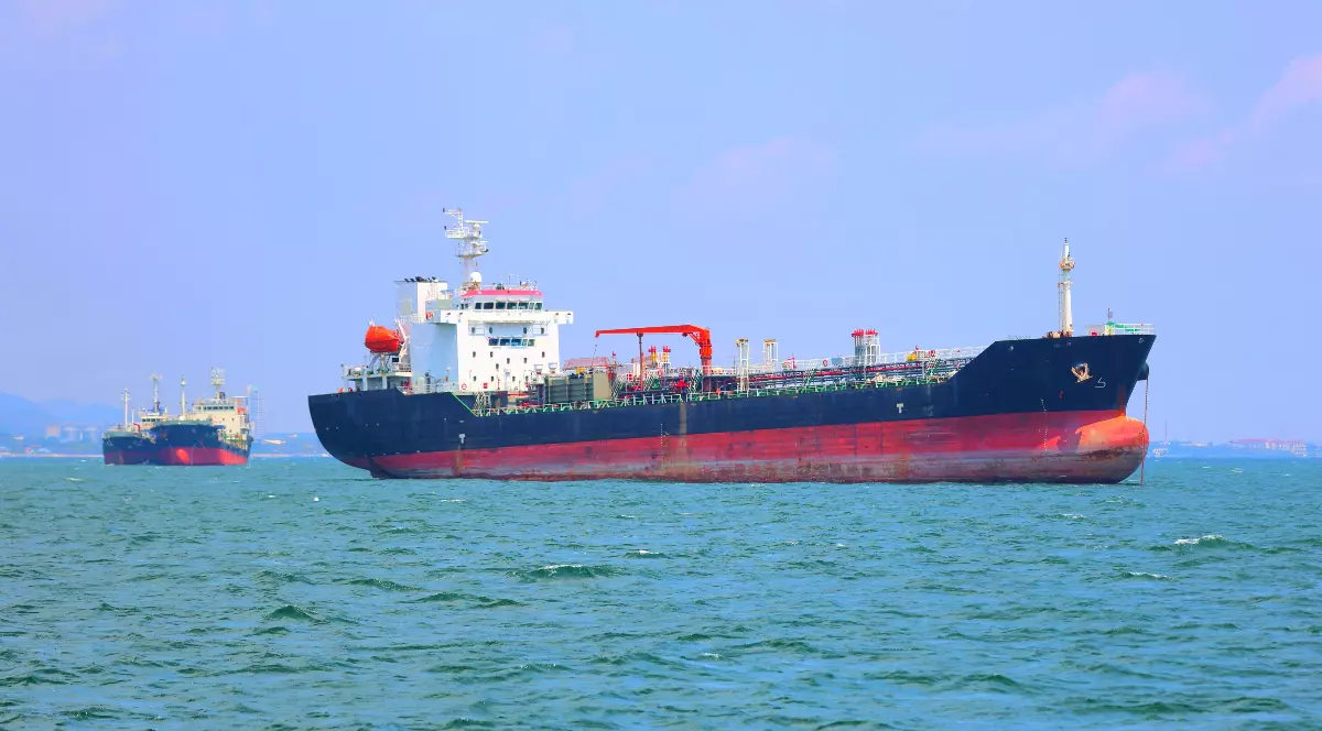Imaginea surprinde un petrolier de mari dimensiuni care navighează pe mare, având în fundal alte nave similare. Cerul este senin, iar scena sugerează o zonă intens circulată pentru transportul maritim de resurse.