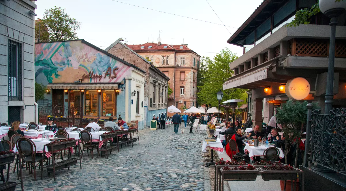 O stradă pietruită, plină de farmec, este mărginită de clădiri vechi și restaurante cochete, unde oamenii stau la mese în aer liber și se bucură de atmosferă. Luminile calde și decorul rustic creează un aer boem, amintind de vremuri mai liniștite și de tradițiile locului.
