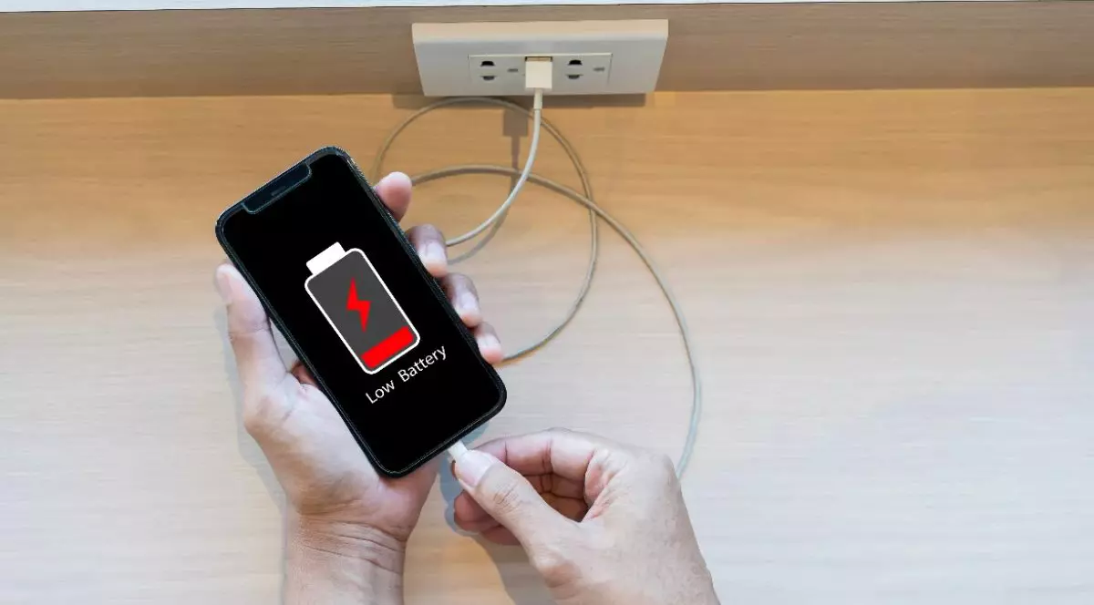 Imaginea arată o persoană care conectează un telefon mobil la încărcător, în timp ce pe ecran apare avertizarea de baterie descărcată. Scena sugerează necesitatea reîncărcării dispozitivului și utilizarea frecventă a telefonului în viața de zi cu zi.