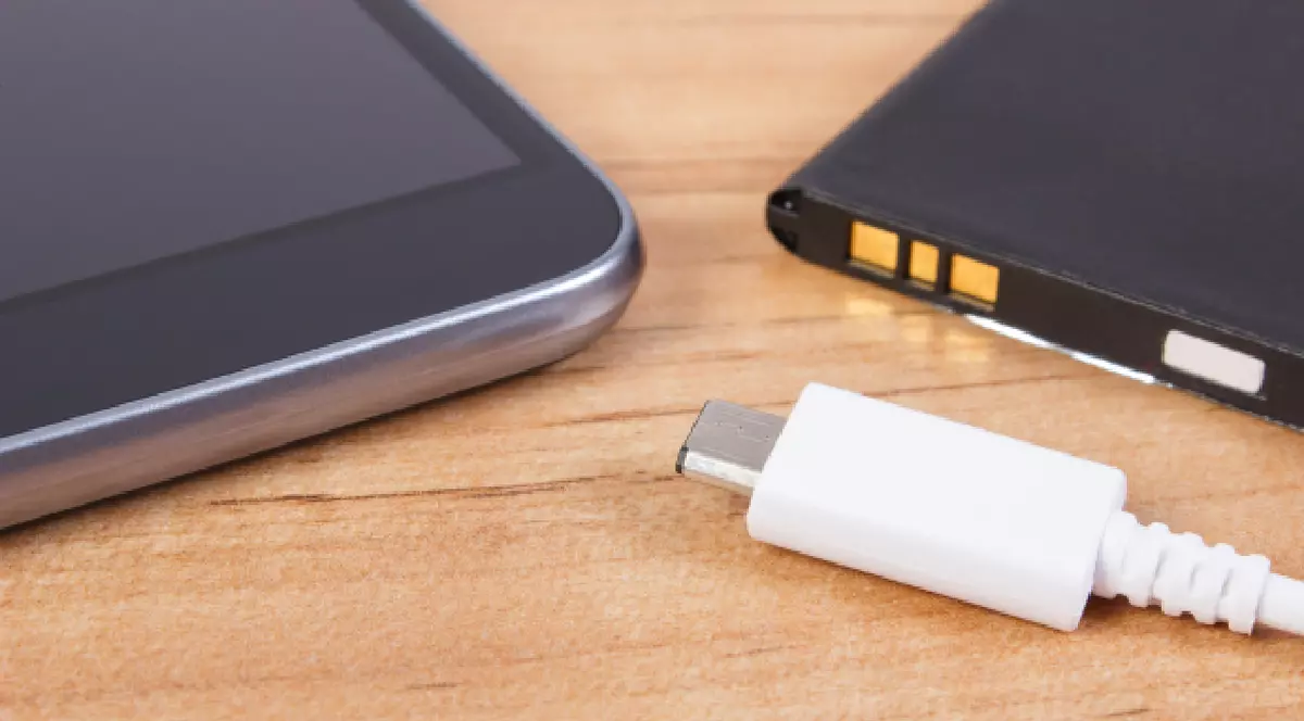 O imagine în prim-plan prezintă un dispozitiv mobil, o baterie și un cablu de încărcare așezate pe o suprafață cu aspect de lemn.