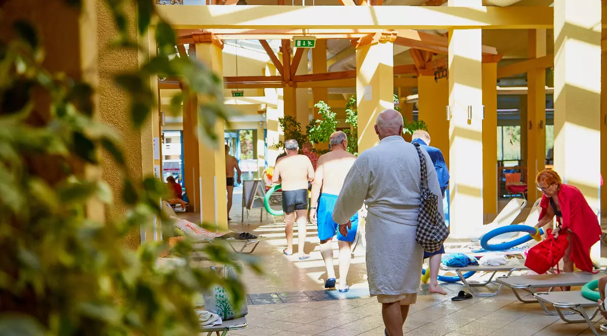 Imaginea surprinde mai multe persoane, în special vârstnici, aflate într-un centru balnear, deplasându-se spre zona piscinelor sau de tratament. Atmosfera este relaxată, cu oameni în halate sau costume de baie, sugerând un cadru dedicat recuperării și odihnei.