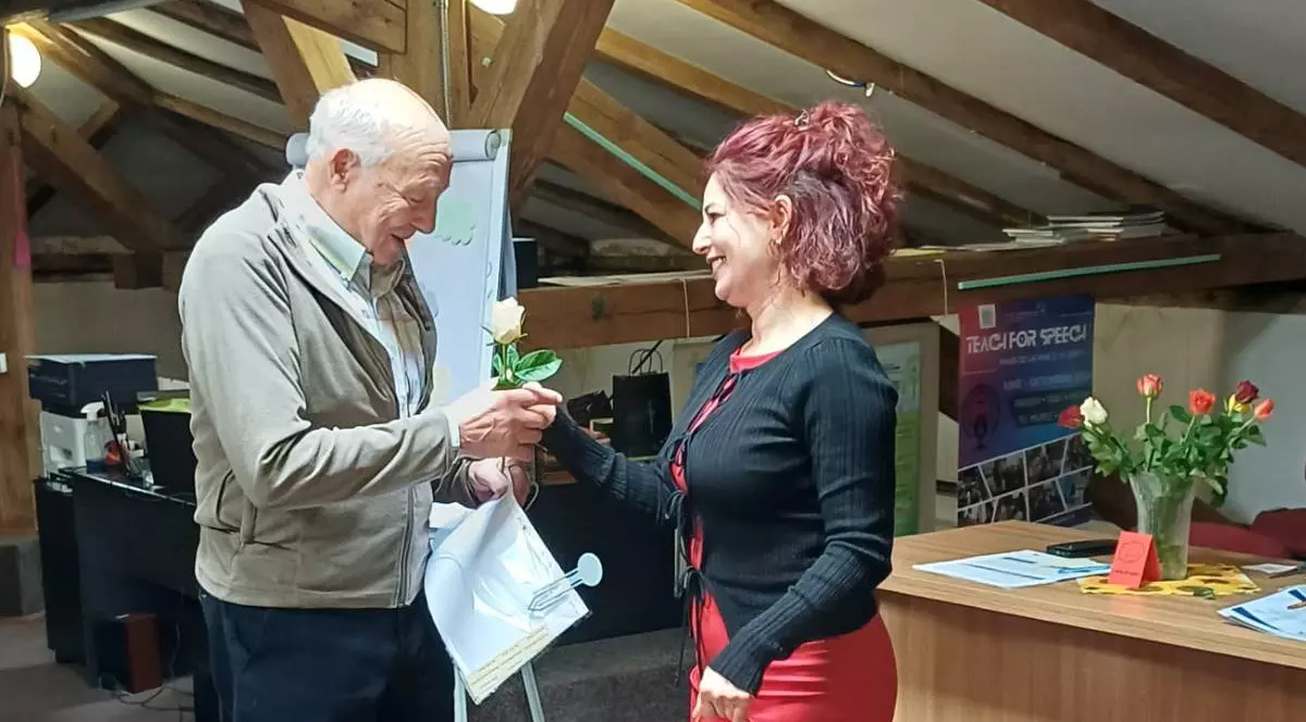 Bărbat în vârstă, Tudor Opincă, într-o încăpere cu grinzi de lemn și primește diploma de absolvire și o floare de la o femeie care zâmbește. 1 Aprilie 2026