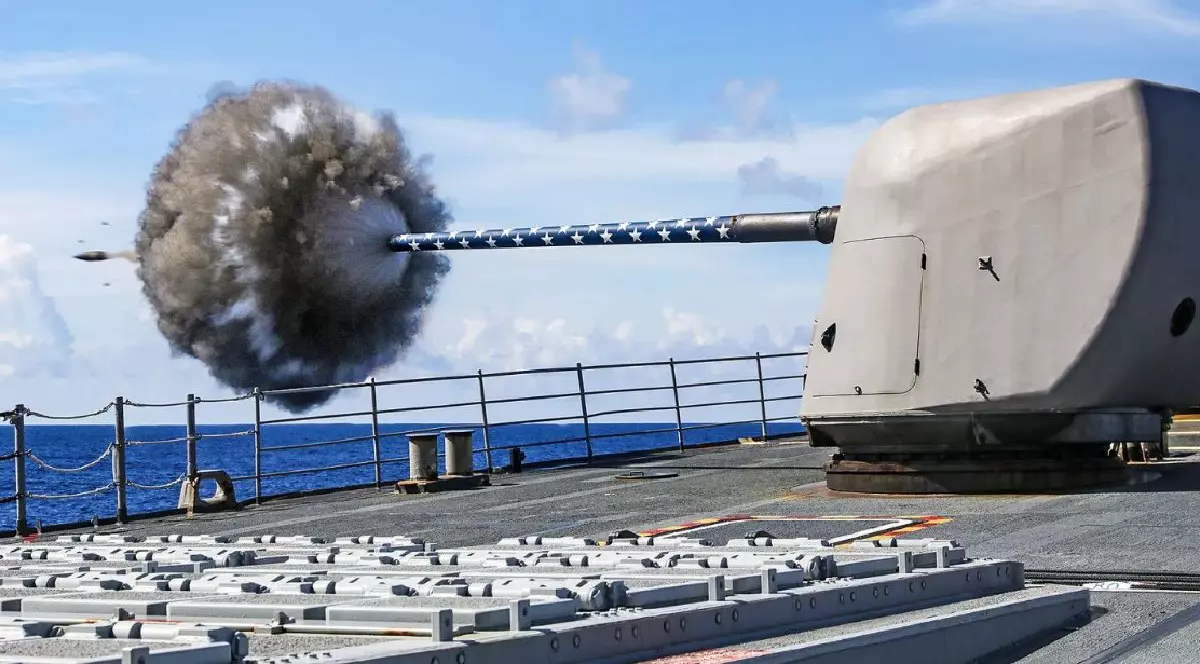 O fotografie de pe puntea unui distrugător surprinde momentul în care un tun Mk 45 Mod 4, având o țeavă decorată cu stele, trage un proiectil, generând o erupție puternică de fum și foc la bot, cu marea deschisă și cerul înstelat în fundal.