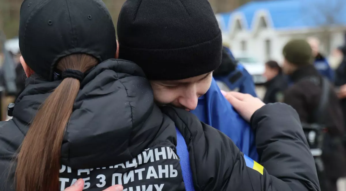 Imaginea surprinde două persoane care se îmbrățișează strâns, într-un moment emoționant, sugerând o revedere sau un rămas-bun. În fundal se observă mai multe persoane, iar pe haine apare un text în limba ucraineană, indicând un context legat de Ucraina.