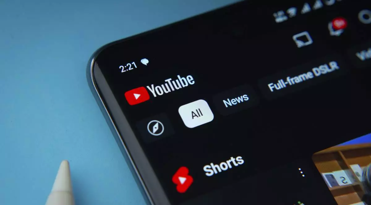 Aplicația YouTube, deschiusă pe ecranul unui smartphone așezat pe un fundal albastru deschis.