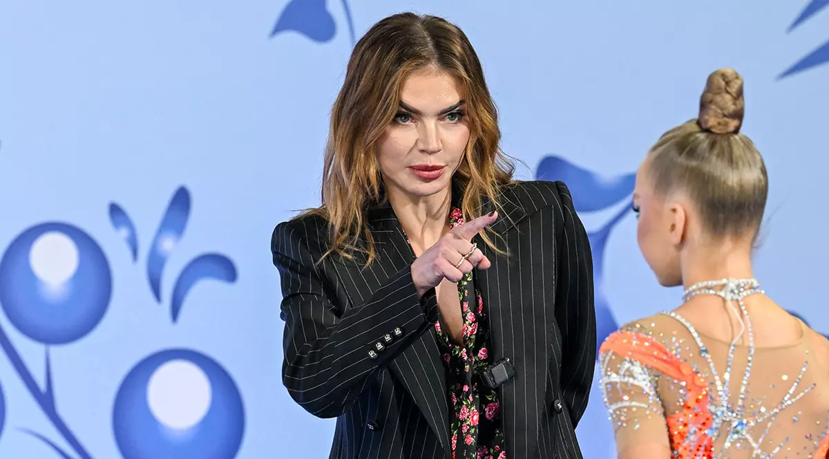 Alina Kabaeva îmbrăcată într-un costum negru discută cu o gimnastă care este cu spatele.