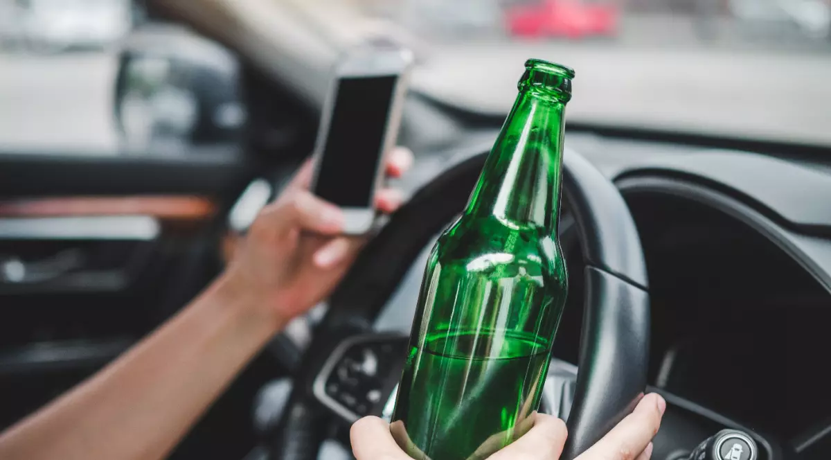 Un bărbat care conduce o mașină, dar într-o mână ține un telefon mobil și în altă mână o sticlă de bere
