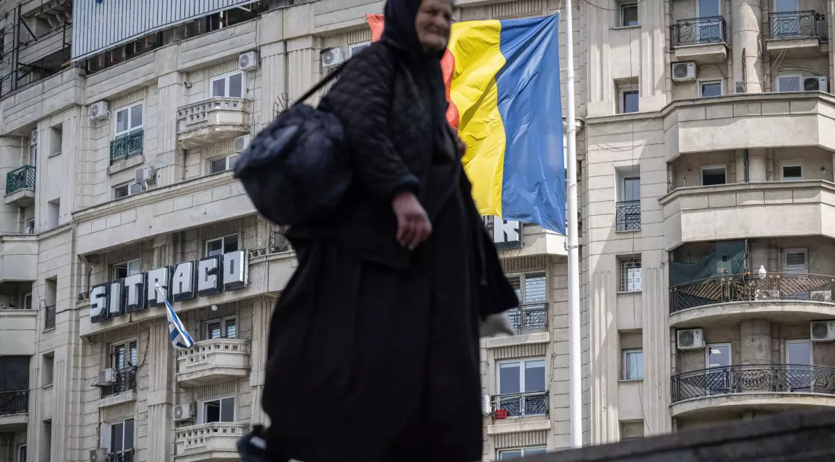 O femeie în vârstă, îmbrăcată în negru și cu batic pe cap, iar pe fundal se vede o clădire și steagul României