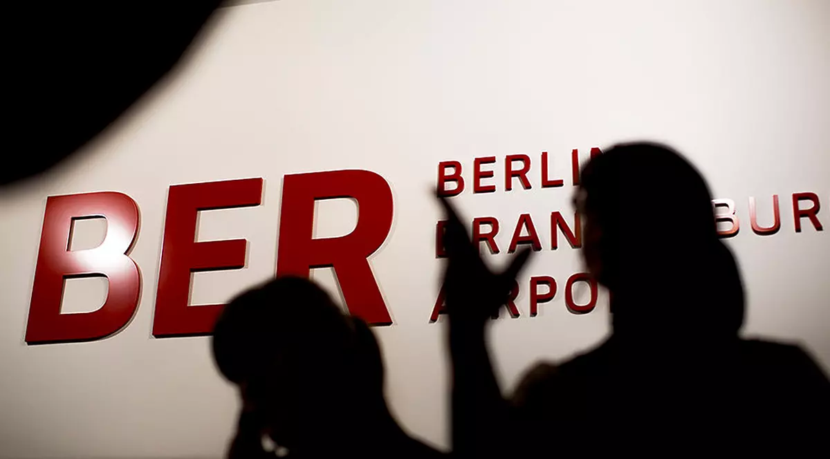 Două persoane in umbră iar în spate se vede sris FLUGHAFEN BERLIN BRANDENBURG, adică Aeroportul Berlin Brandenburg.