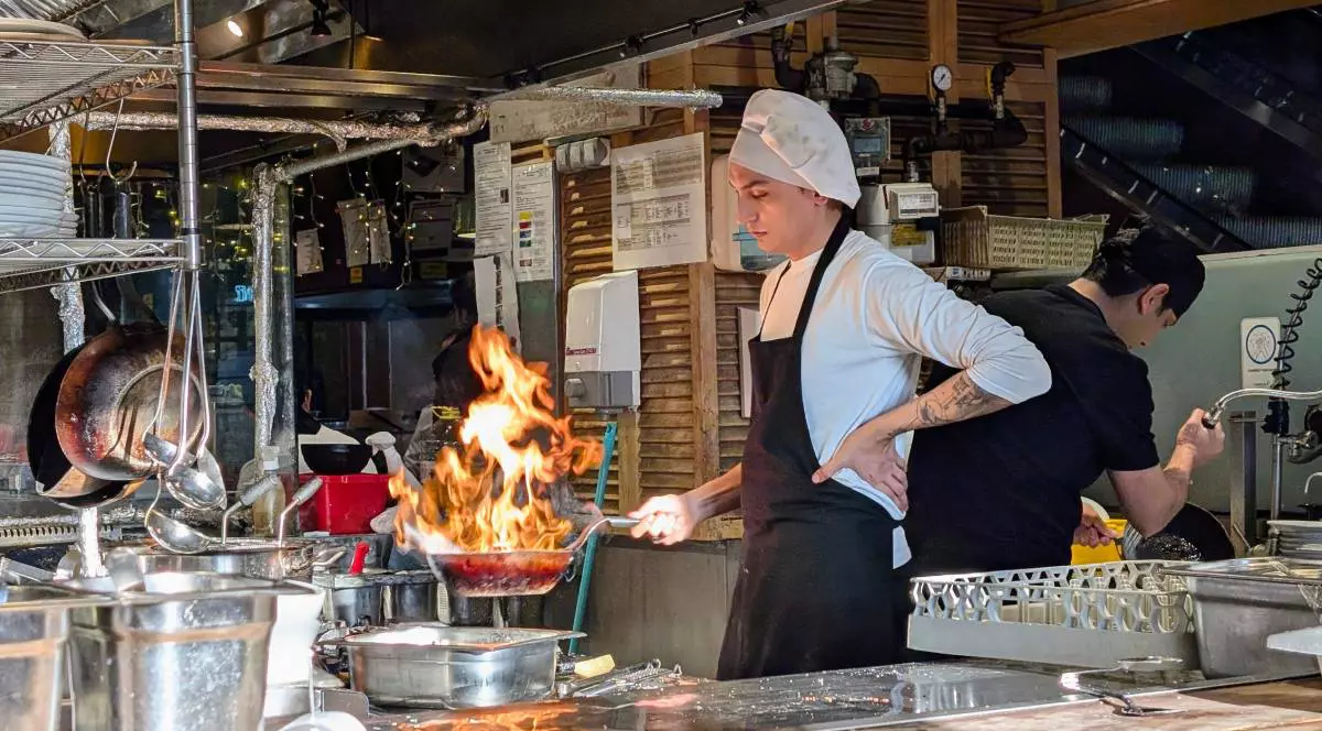 Bucătar cu pălărie albă pregătește un fel de mâncare gourmet flambat într-o tigaie în bucătăria unui restaurant