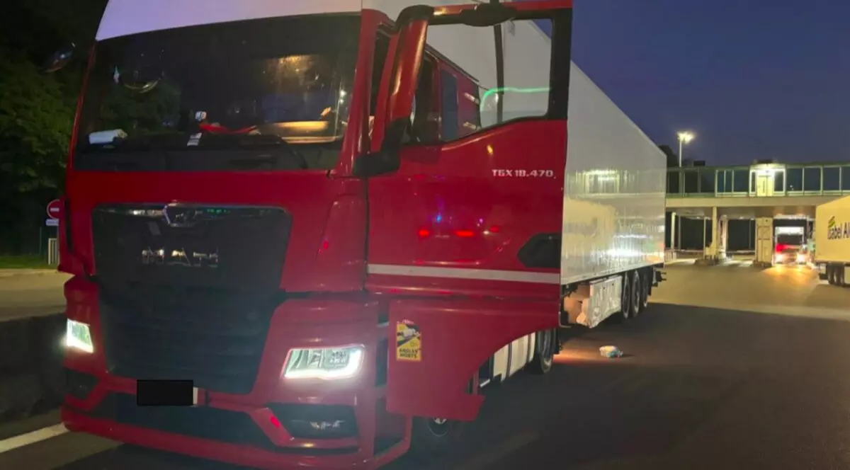 Imaginea prezintă un TIR roșu cu semiremorcă albă, staționat, noaptea, într-o zonă slab iluminată, cu alte camioane în fundal