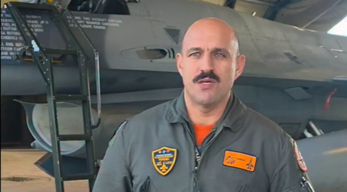 Imaginea îl surprinde pe un pilot militar în uniformă de zbor, aflat în fața unui avion de luptă, într-un hangar sau pe o bază aeriană. Fotografia sugerează un context legat de aviația militară și de misiuni de apărare aeriană.