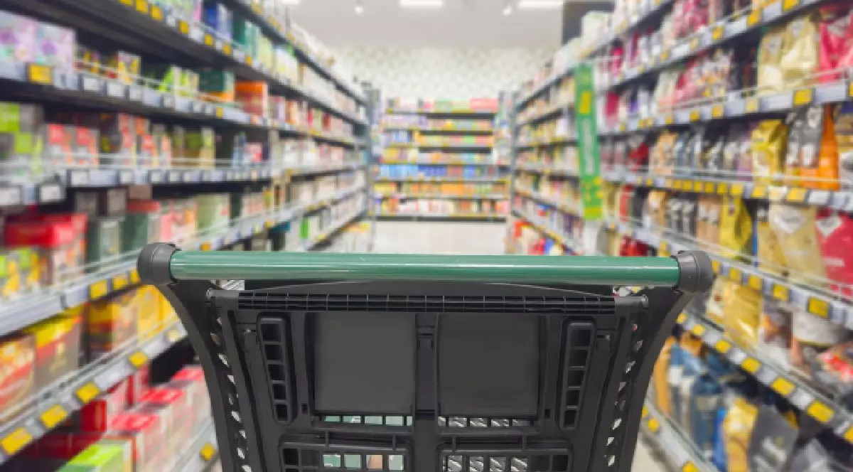 Prim-plan cu un cărucior de cumpărături, pe culoarul unui supermarket, cu cu raftru de o parte și de alta