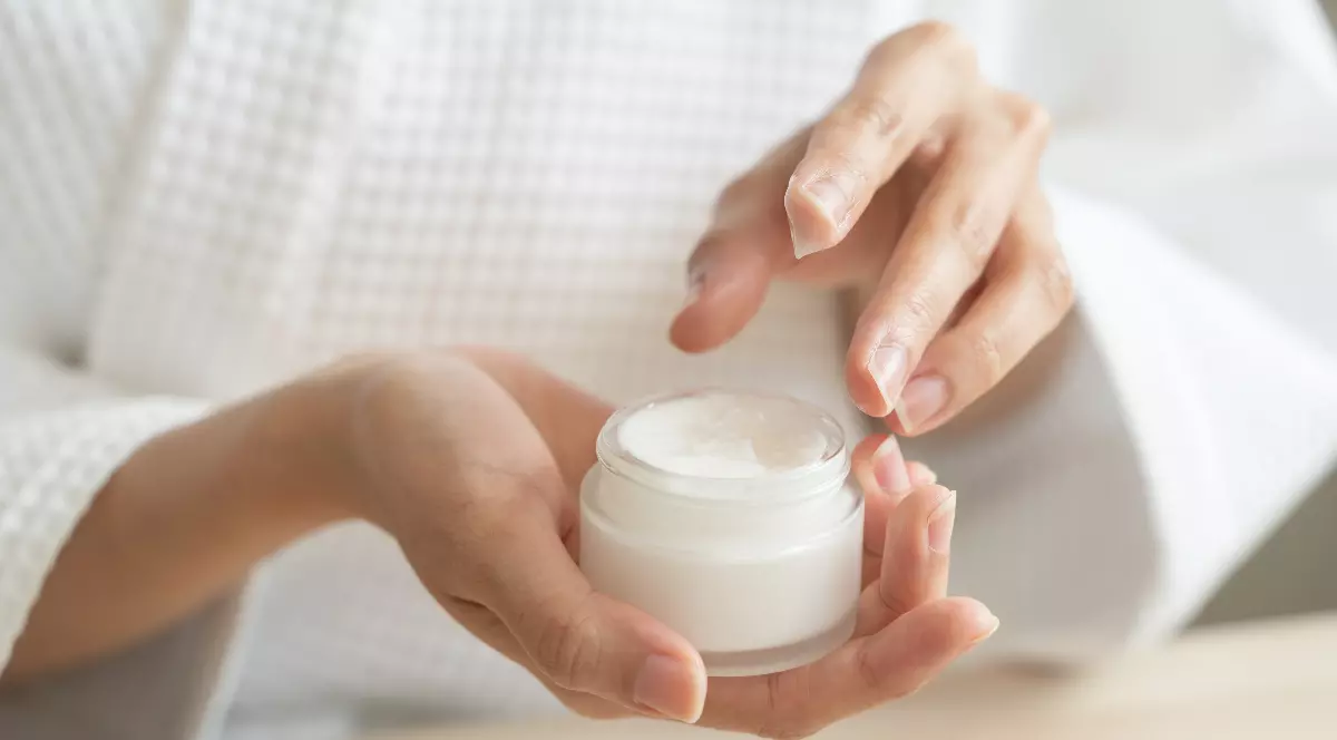 O persoană ține în palmă un recipient cu cremă cosmetică și se pregătește să aplice produsul pe piele. Imaginea sugerează o rutină de îngrijire personală și evidențiază utilizarea unui produs destinat hidratării și protecției pielii.