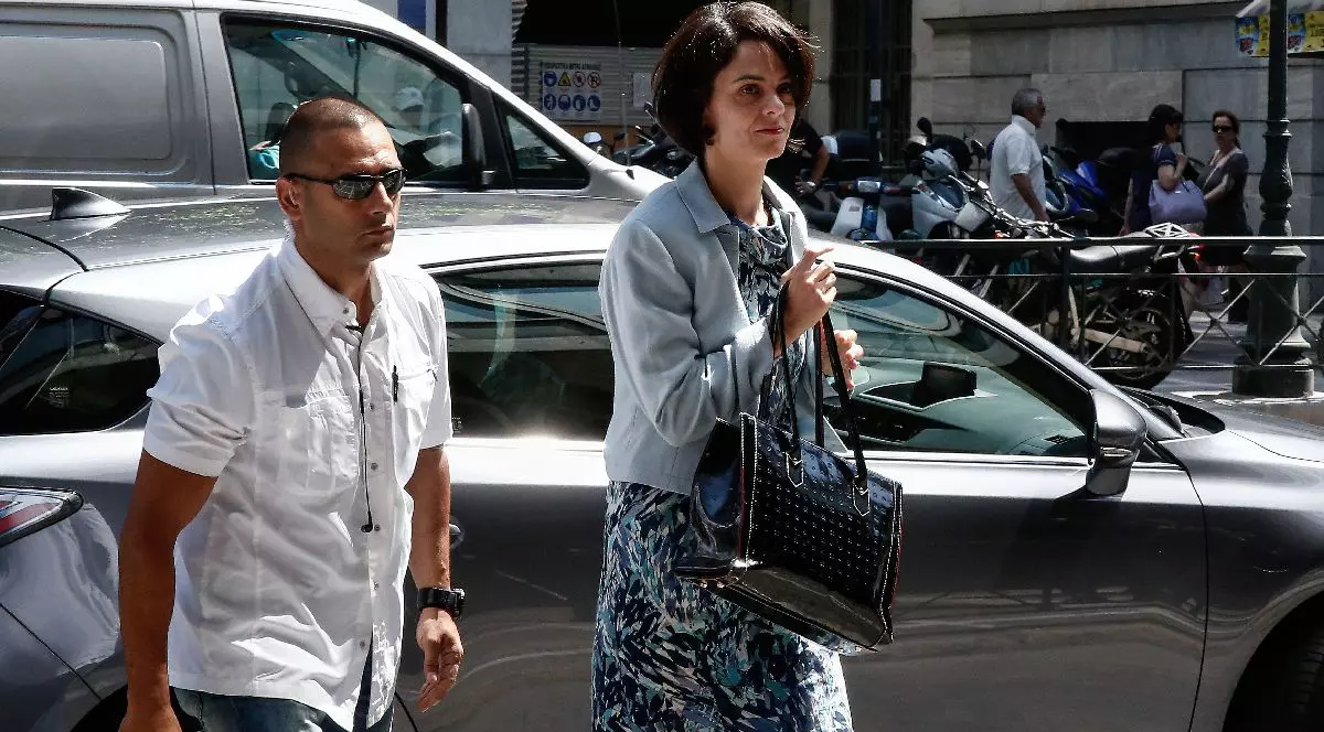 O femeie tunsă scurt cu o geantă în mână, pe o stradă din Atena, în spatele ei un bărbat cu cămașă albă