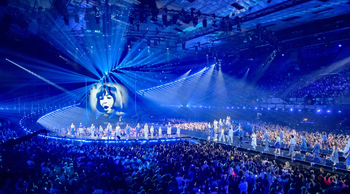 Imaginea surprinde atmosfera spectaculoasă a unui show Eurovision, cu scenă luminată intens și mii de spectatori prezenți în arenă. Efectele vizuale și energia publicului evidențiază amploarea unuia dintre cele mai urmărite concursuri muzicale din lume.