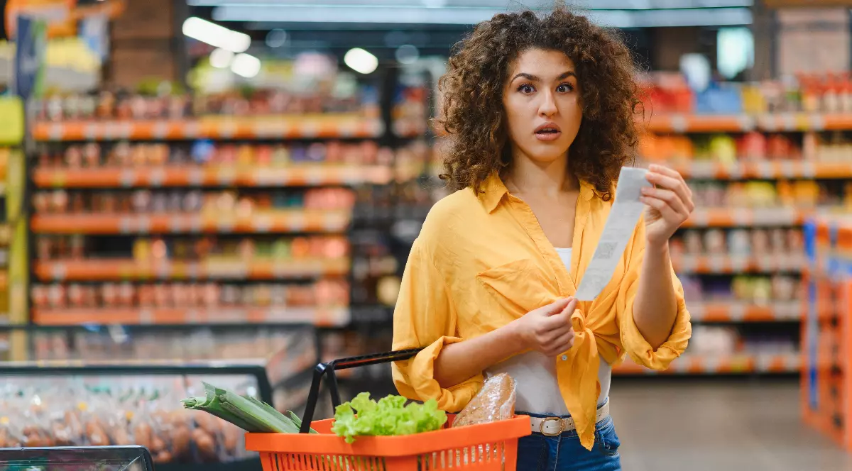 O tânără aflată într-un supermarket privește surprinsă un bon fiscal, în timp ce ține în mână un coș cu produse alimentare. Expresia ei de uimire sugerează că valoarea cumpărăturilor sau prețurile afișate pe bon au luat-o prin surprindere.