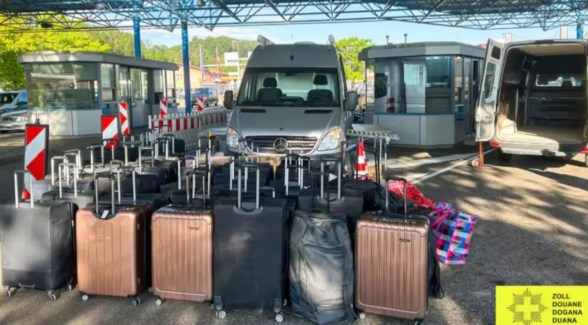 55 de valize și trollere așezate în parcarea unui aeroport, în spatele lor sunt parcate microbuze