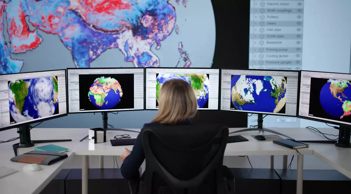Imaginea prezintă o persoană văzută din spate care monitorizează hărți meteorologice și imagini ale globului pământesc afișate pe mai multe monitoare mari, într-un centru de comandă sau analiză a datelor.